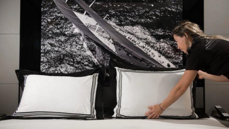 Kiralık lüks bir yatın yatağını hostes düzeltiyor yatak başlığındaki tekne görseli yastıklarla uyumlu.